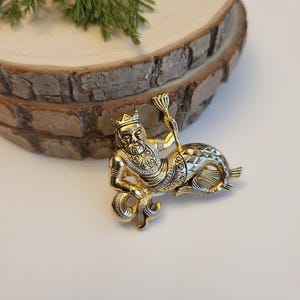 May include: A gold-coloured brooch featuring a mythical figure with a human upper body wearing a crown and holding a staff. The lower body is a fish tail. The brooch has detailed patterns and is on a wooden slice.