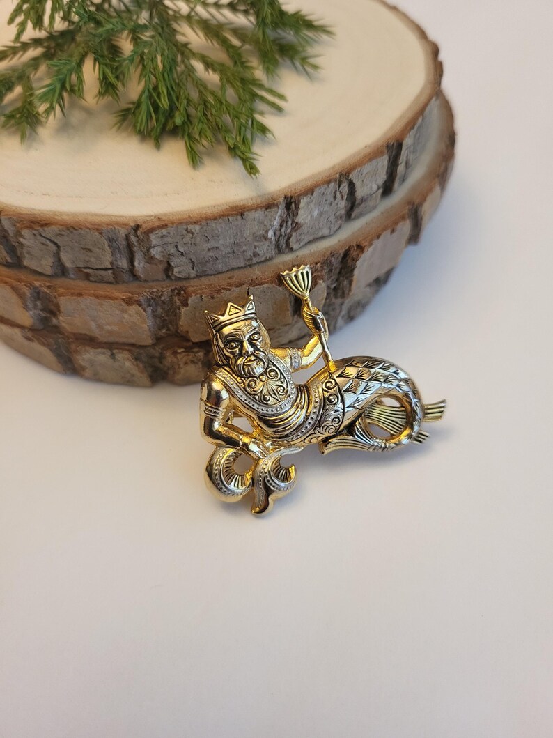 May include: A gold-coloured brooch featuring a mythical figure with a human upper body wearing a crown and holding a staff. The lower body is a fish tail. The brooch has detailed patterns and is on a wooden slice.
