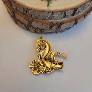 May include: A gold-coloured vintage brooch depicting a mythical creature with a horn and a trumpet. The brooch has detailed features and a pin fastening on the reverse. The brooch is set against a light background with a wooden slice and greenery.
