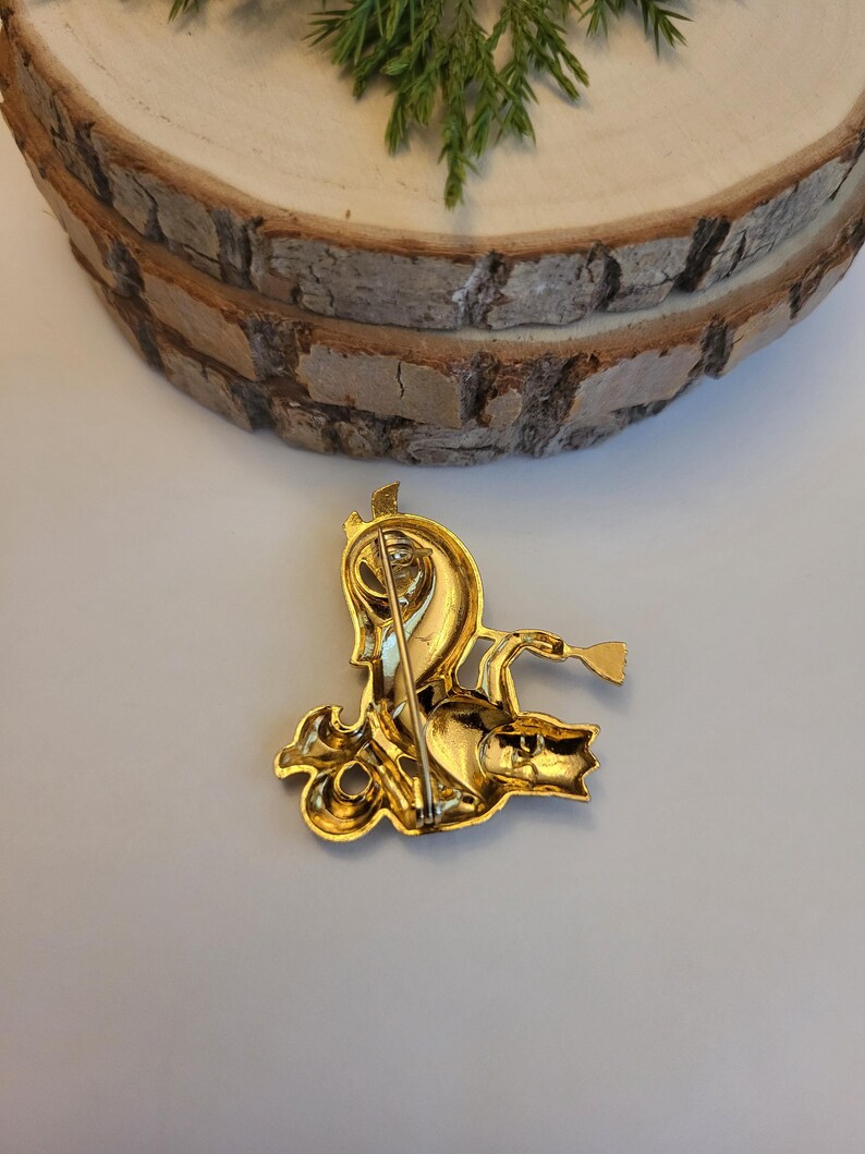 May include: A gold-coloured vintage brooch depicting a mythical creature with a horn and a trumpet. The brooch has detailed features and a pin fastening on the reverse. The brooch is set against a light background with a wooden slice and greenery.