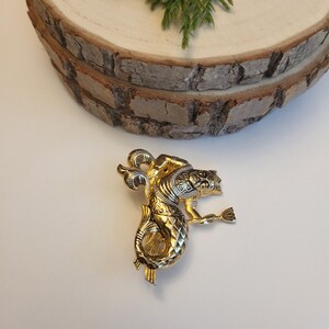 May include: A gold-coloured metal brooch depicting a mythical creature, possibly a seahorse or dragon. The brooch features detailed craftsmanship, with gold and silver tones. The creature is shown with a flowing tail and an outstretched arm.