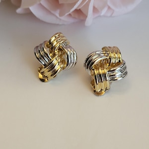 May include: A pair of clip-on earrings featuring a knotted design with alternating polished gold-tone and silver-tone metal bands. The earrings have a rounded, dimensional shape with a smooth, reflective finish on each of the interlocking metal loops.