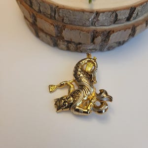 May include: A gold brooch with an elaborate design, depicting a figure playing a horn. The brooch has detailed patterns and a shiny finish. The background includes a wooden slice and some greenery.