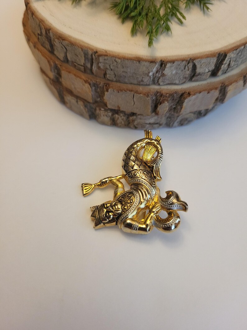 May include: A gold brooch with an elaborate design, depicting a figure playing a horn. The brooch has detailed patterns and a shiny finish. The background includes a wooden slice and some greenery.