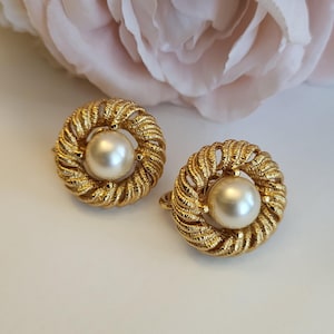 May include: A pair of gold-toned clip-on earrings. Each earring features a large, round, cream-colored faux pearl at the center, surrounded by a textured, woven gold metal design. The earrings are set against a soft, white background.
