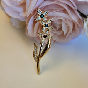 May include: A gold-toned floral brooch featuring three flower blossoms with jewel-toned centers and clear rhinestone accents. The brooch has a stem and leaf design, perfect for adding a touch of elegance to any outfit. The brooch is set against a backdrop of pink flowers.