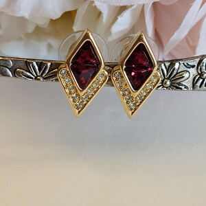 May include: A pair of gold-tone stud earrings featuring a diamond-shaped design. Each earring contains a deep red faceted stone at the top and a row of small clear crystals along the bottom edges, secured with clear plastic earring backs.