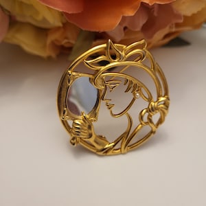 May include: A gold-toned vintage brooch featuring a woman looking into a mirror. The brooch has an Art Nouveau design with a woman's profile, a pearl earring, and decorative flourishes. The brooch is round and has a polished finish.