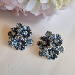 May include: A pair of blue flower-shaped earrings with silver-colored metal accents. Each earring features multiple flower clusters with light blue crystal-like embellishments in the center and around the petals. The earrings are set against a soft white background.