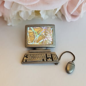 May include: A pewter-toned metal pin shaped like a vintage desktop computer with a keyboard and a separate mouse attached by a thin chain. The monitor screen features a holographic insert with a pattern of intersecting light-colored lines.