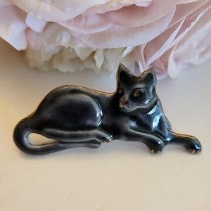 May include: A small, glossy black ceramic cat figurine in a reclining position. The cat has a smooth, dark finish with subtle gold-toned metallic edging along its ears, back, and paws, highlighting its sleek, elongated form.