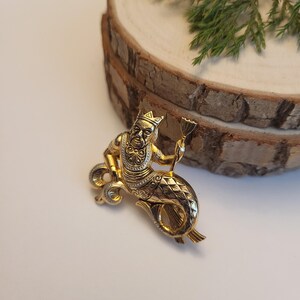 May include: A gold-coloured brooch featuring a mythical figure with a human upper body and a fish tail. The figure wears a crown and holds a small item. The brooch has detailed patterns and is set against a plain background.