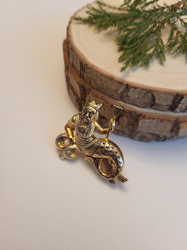 May include: A gold-coloured brooch featuring a mythical figure with a human upper body and a fish tail. The figure wears a crown and holds a small item. The brooch has detailed patterns and is set against a plain background.