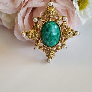 May include: A gold-toned vintage brooch with an oval, marbled green stone center. The brooch features ornate detailing, including lion head accents and small faux pearls. The background includes pink and white flowers.