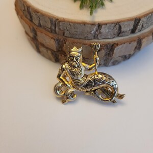 May include: A gold-coloured brooch featuring a mythical sea creature wearing a crown and holding a chalice. The brooch has detailed features on the face, beard, and fish tail. The brooch is set against a white background.