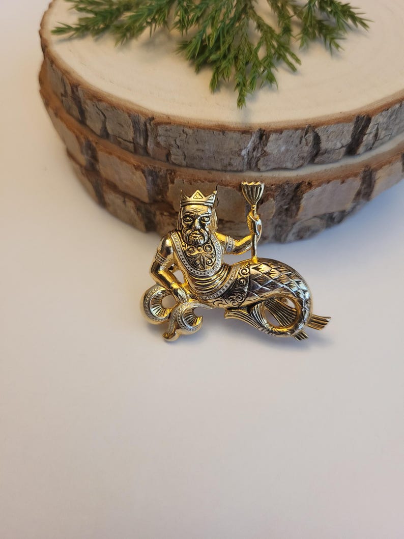 May include: A gold-coloured brooch featuring a mythical sea creature wearing a crown and holding a chalice. The brooch has detailed features on the face, beard, and fish tail. The brooch is set against a white background.