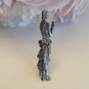 May include: A silver-toned metal brooch shaped like a standing figure wearing a dress with a bow detail at the waist. The surface of the dress is embellished with small, dark, faceted stones that catch the light, creating a textured appearance.