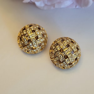May include: A pair of gold-toned, round clip-on earrings. Each earring features a woven basket design encrusted with small, sparkling crystals. The earrings are set against a soft, neutral background, with a hint of a pink flower in the upper corner.