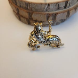 May include: A gold-coloured metal brooch featuring a mythical sea king wearing a crown and holding a chalice. The king has a detailed face and is seated on a fish-like tail. The brooch has intricate detailing and a vintage aesthetic.