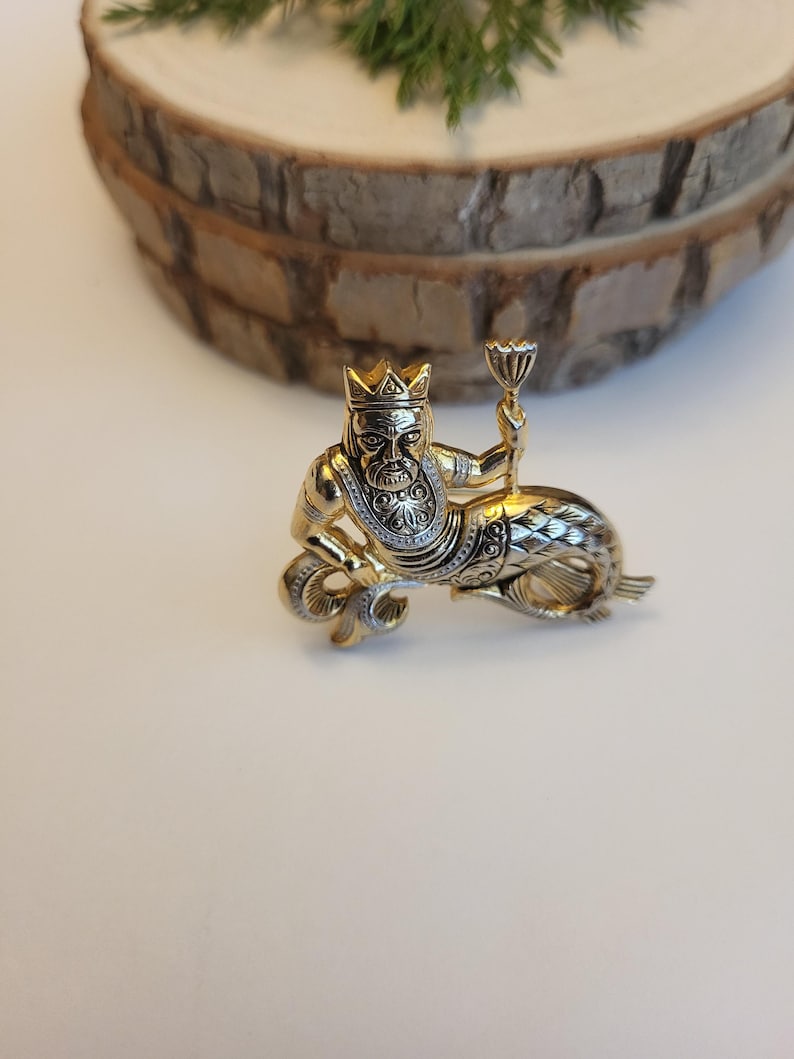 May include: A gold-coloured metal brooch featuring a mythical sea king wearing a crown and holding a chalice. The king has a detailed face and is seated on a fish-like tail. The brooch has intricate detailing and a vintage aesthetic.