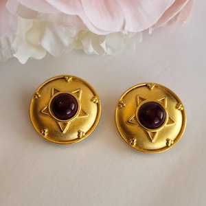 May include: A pair of gold-toned, round earrings. Each earring features a six-pointed star design with a dark red, circular stone in the center. The earrings have a vintage aesthetic and are set against a soft, neutral background.