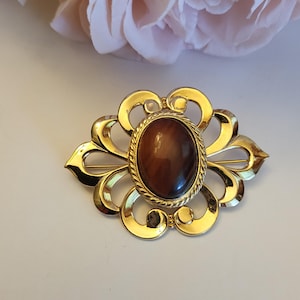 May include: A gold-toned brooch with an oval, brown stone center. The brooch has a decorative, openwork design with a floral shape. The stone is surrounded by a twisted rope-like border. The brooch is likely vintage or antique.