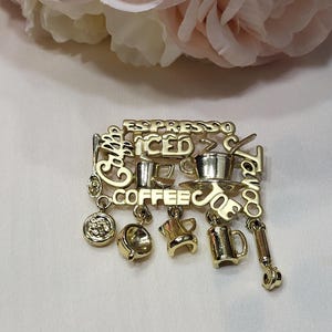 May include: Gold-toned coffee-themed brooch with the words "Espresso", "Iced", and "Coffee" in a stylized font. The brooch features miniature coffee cups, a coffee pot, and other coffee-related charms.