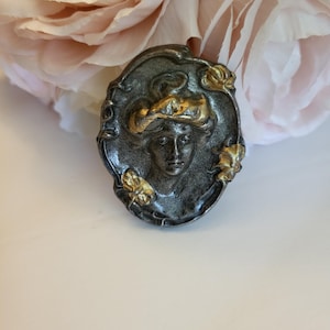 May include: An oval-shaped brooch featuring a relief portrait of a person with wavy hair, rendered in a dark metallic gray finish with gold-toned accents on the hair and floral elements along the ornate, textured border of the frame.