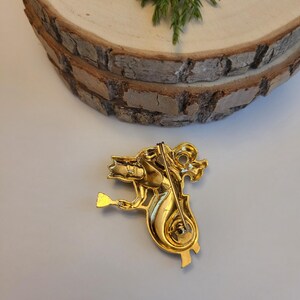 May include: A gold-coloured brooch depicting a seahorse playing a stringed instrument. The brooch features intricate detailing and a pin fastening. The seahorse is facing left, holding the instrument with its front flipper.