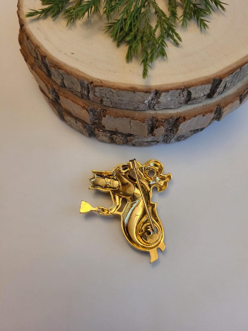 May include: A gold-coloured brooch depicting a seahorse playing a stringed instrument. The brooch features intricate detailing and a pin fastening. The seahorse is facing left, holding the instrument with its front flipper.