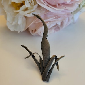 May include: A metallic brooch in the shape of a long-necked bird standing among stylized reeds. The piece features a dark, textured bronze-toned finish with a slender, upright neck and curved, pointed elements representing foliage at the base.