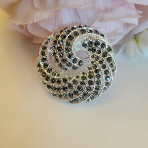 May include: Silver-toned brooch with a spiral design, encrusted with dark gray rhinestones. The brooch has a pin closure on the back. The background features a soft pink flower.