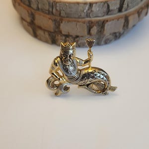 May include: A gold-coloured metal brooch featuring a crowned figure holding a chalice, atop a fish-like body. The brooch has detailed patterns and a two-tone finish, with silver accents. The background is a light surface with a wooden slice.