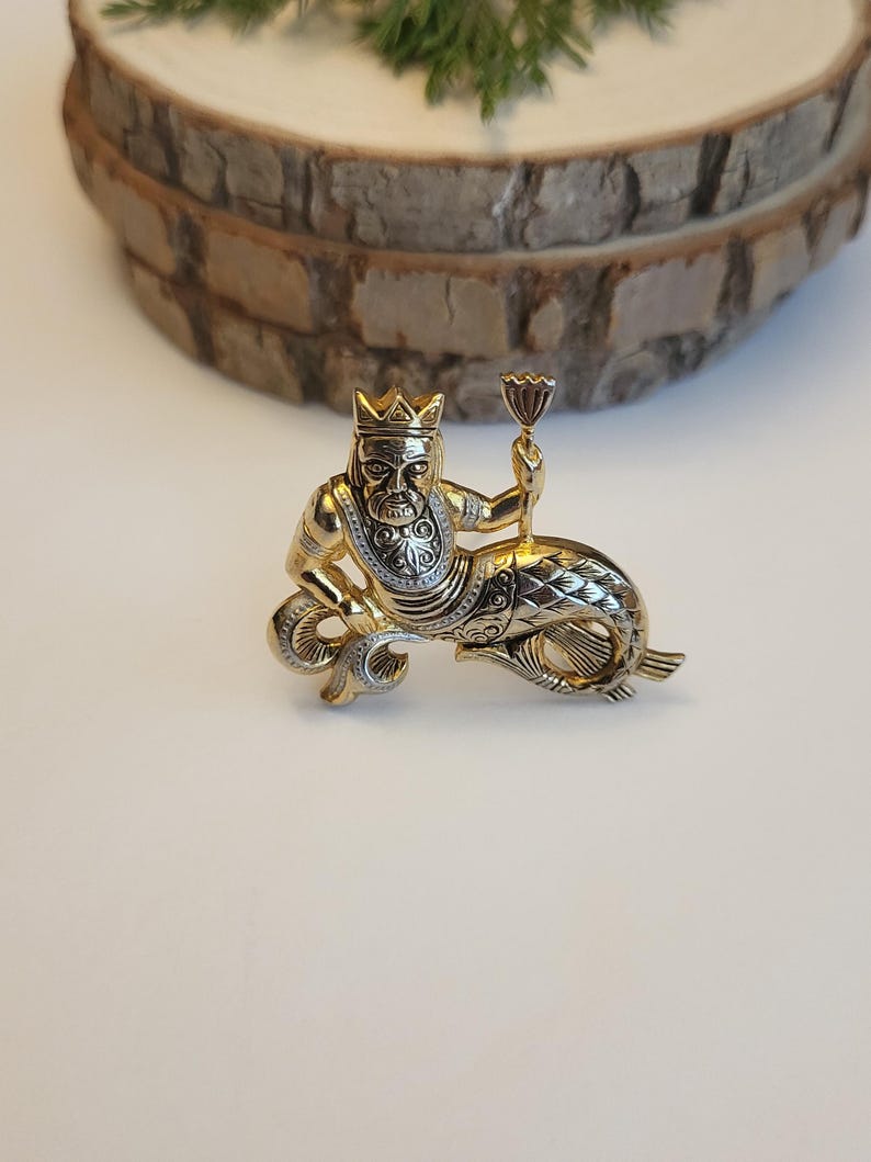 May include: A gold-coloured metal brooch featuring a crowned figure holding a chalice, atop a fish-like body. The brooch has detailed patterns and a two-tone finish, with silver accents. The background is a light surface with a wooden slice.