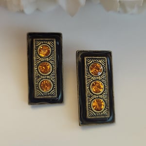 May include: A pair of rectangular earrings featuring a black enamel border surrounding a central antique gold-toned panel. Each earring is decorated with three round, faceted amber-colored crystals arranged vertically in a row.