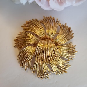 May include: A gold-tone brooch shaped like a flower with multiple layered petals. Each petal features a textured, rope-like finish that radiates outward from the center, creating a dense, circular, and dimensional floral design.