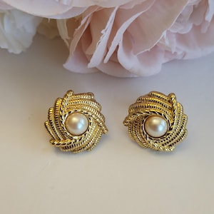 May include: A pair of gold-toned clip-on earrings. Each earring features a central faux pearl surrounded by a woven, rope-like design. The earrings are set against a soft, blurred background with a pink flower.