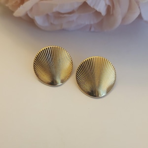 May include: Two gold-colored, round earrings with a ridged, fan-like design. The earrings are set against a soft, off-white background with a blurred pink flower in the upper corner. These earrings are a classic jewelry accessory.