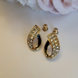 May include: A pair of gold-toned stud earrings with a teardrop design. The earrings feature clear crystals and a black enamel accent. The earring posts are gold-colored. The background is a soft white.