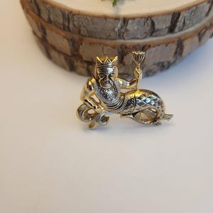 May include: A gold-coloured brooch featuring a mythical sea creature with a crown, holding a chalice. The figure has a human upper body and a fish tail, with detailed silver accents. The brooch is set against a white surface.