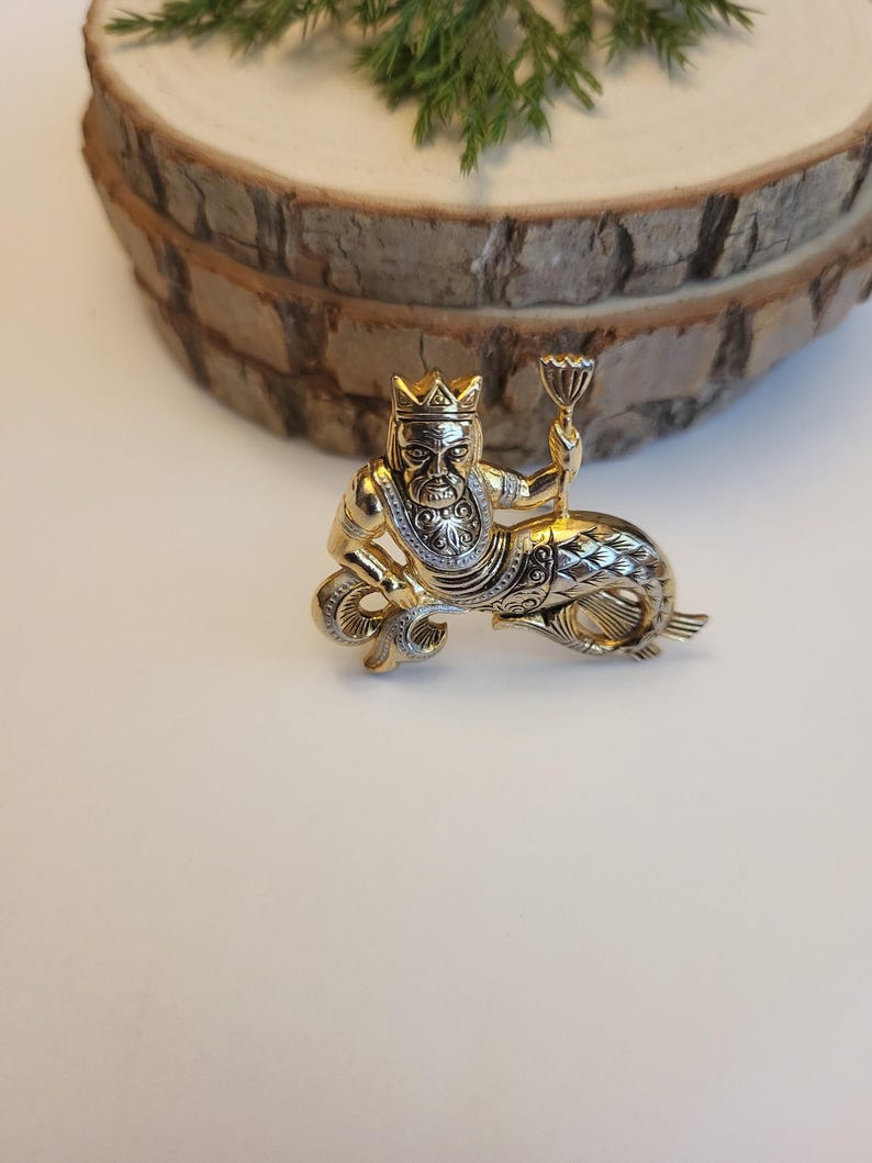 May include: A gold-coloured brooch featuring a mythical sea creature with a crown, holding a chalice. The figure has a human upper body and a fish tail, with detailed silver accents. The brooch is set against a white surface.