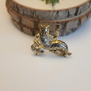 May include: A gold-coloured brooch depicting a mythical king figure with a crown and sceptre, riding a fish. The brooch has detailed craftsmanship and a regal design. The background is neutral.