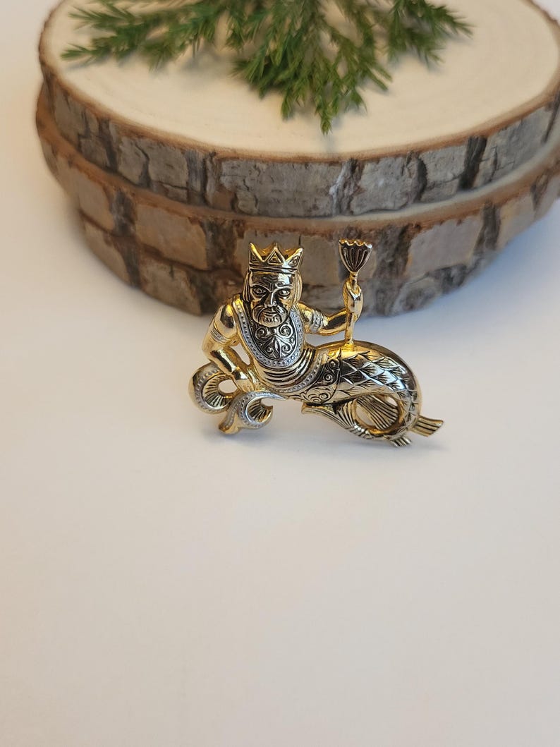 May include: A gold-coloured brooch depicting a mythical king figure with a crown and sceptre, riding a fish. The brooch has detailed craftsmanship and a regal design. The background is neutral.