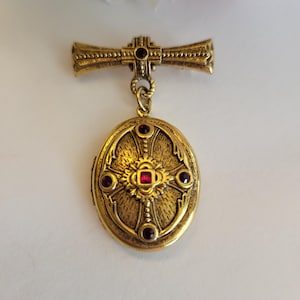May include: A gold-toned metal brooch featuring a horizontal bar top piece with a small red stone, connected by a ring to an oval locket pendant. The locket has a textured surface, a central red stone, and four smaller red stones in a cross pattern.