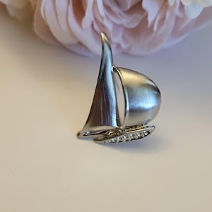 May include: Silver-toned sailboat brooch with a curved sail and a row of clear rhinestones along the bottom. The brooch has a polished finish and a nautical theme. The background features soft pink and white flowers.
