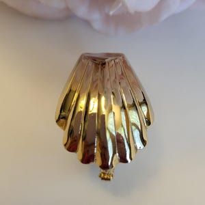 May include: A gold-toned metal earring shaped like a scallop shell with a polished, reflective surface and vertical ridges. The earring features a clip-on mechanism at the base and a curved, fan-like silhouette that mimics the natural form of a seashell.