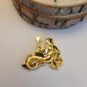 May include: A gold brooch featuring a mermaid-like character riding a seahorse and holding a goblet. The brooch has a pin fastening on the back. The design is detailed and ornate, with a shiny finish.
