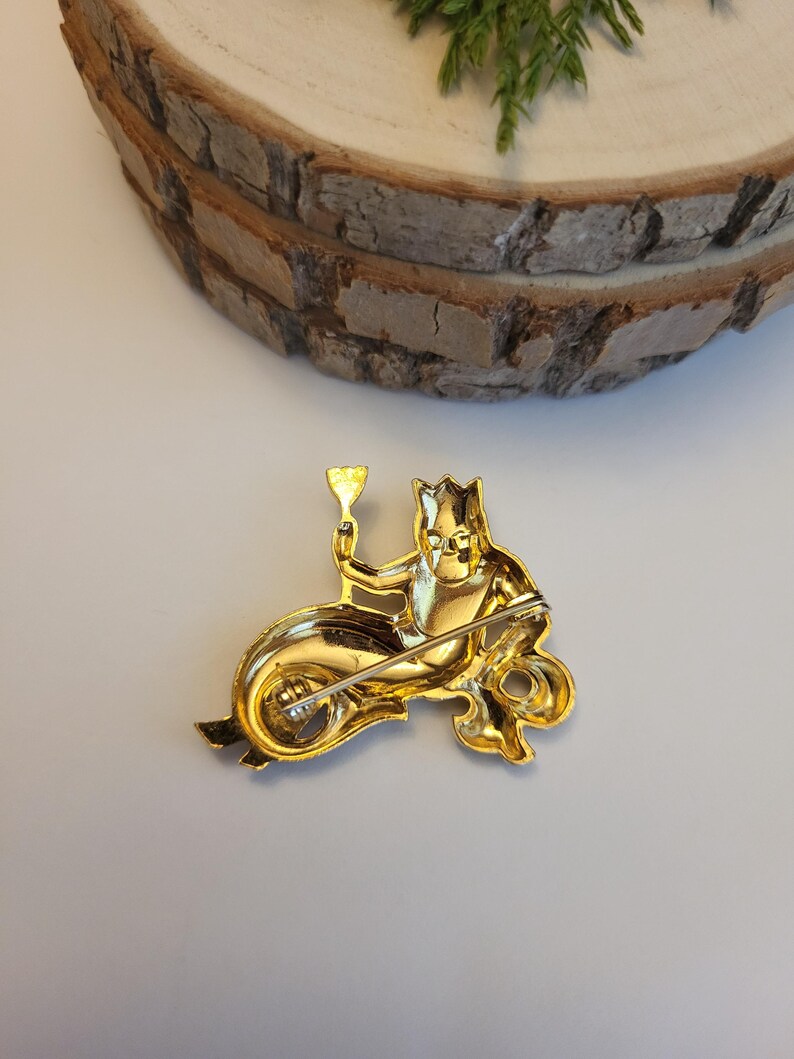 May include: A gold brooch featuring a mermaid-like character riding a seahorse and holding a goblet. The brooch has a pin fastening on the back. The design is detailed and ornate, with a shiny finish.