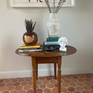 May include: A wooden drop-leaf table with four turned legs, holding a stack of books, a white patterned vase with branches, a dark brown spherical vase with feathers, a small black ceramic bowl, and a white sculpted head bust.