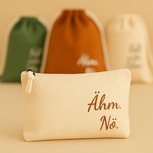 May include: A cream-colored canvas zippered pouch with the words "Ähm. Nö." printed in brown. Three drawstring bags in green, orange, and cream are blurred in the background.